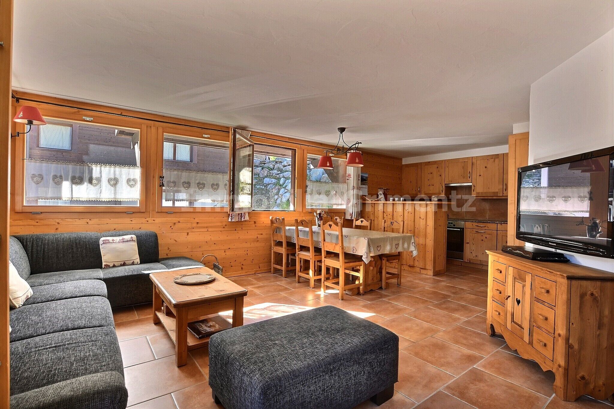 A cozy living room with a sofa, ottoman, and dining table. There is a kitchen area with wooden cabinets and a television.