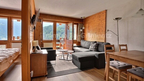 A cozy room with a sofa, a dining table, and a bed. Large windows offer a view of the outdoors. - Comfortable flat for 4 people, living room, kitchen area, 1 2-bedroom, sleeping corner with 1 bunk b (Grimentz)