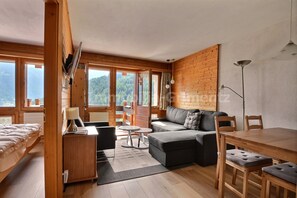 A cozy room with a sofa, a dining table, and a bed. Large windows offer a view of the outdoors. - Comfortable flat for 4 people, living room, kitchen area, 1 2-bedroom, sleeping corner with 1 bunk b (Grimentz)