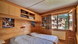 A wooden cabin room with two beds, a window with a view of a mountain village, and wooden shelves with items. - Comfortable flat for 6 people, living room with fireplace, kitchen area with dishwasher, 1 2-bedroom (Grimentz)