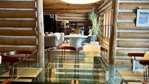 Ein gemütliches Blockhaus-Interieur mit einem Glastisch, um den herum Stühle, eine Couch und eine Lampe stehen.