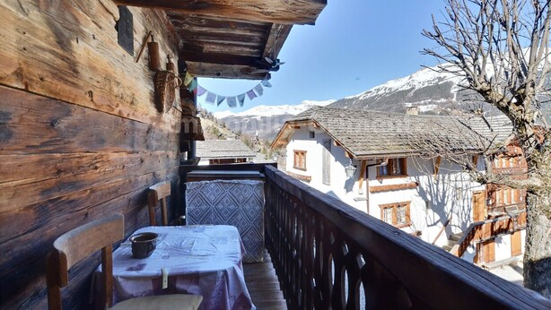 Un balcon en bois, agrémenté d’une table et de chaises, qui offre une vue sur un village de montagne aux bâtiments traditionnels.