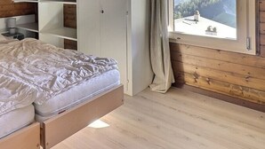 Uma cama de casal com cabeceira de madeira, uma mesa de cabeceira com abajur e vista para o exterior pela janela.