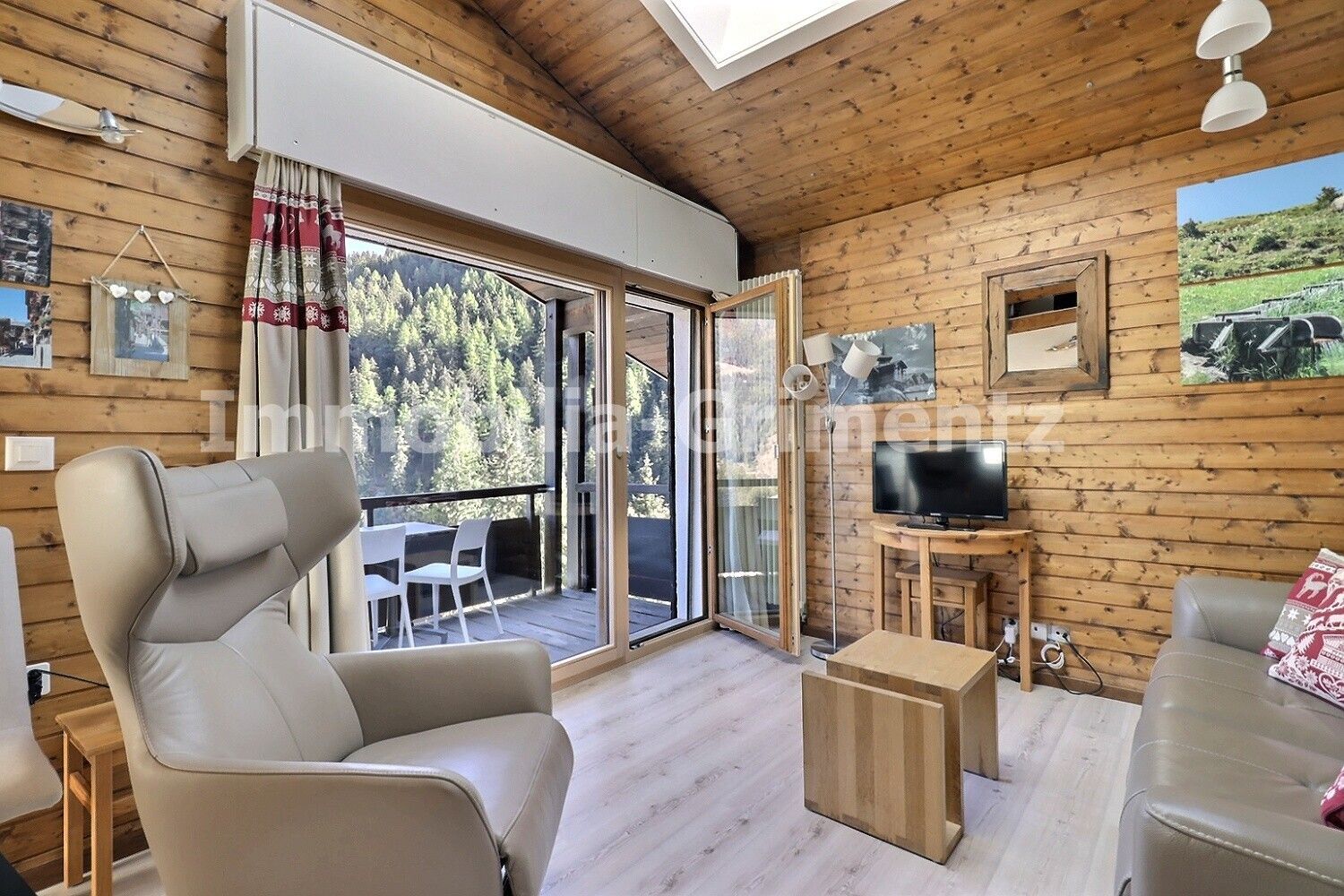 A wooden cabin interior with a sofa, a chair, a television, and a balcony with a view of trees.
