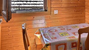 A wooden cabin room with a table covered in a patterned tablecloth, a chair, and a window with curtains.