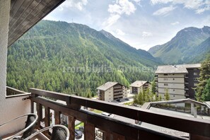 Un balcon offrant une vue sur une chaîne de montagnes boisée, un immeuble à plusieurs étages et un ciel dégagé.