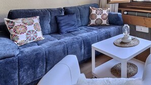 Ett vardagsrum med en blå soffa, vita fåtöljer, ett litet vitt bord med en dekorativ detalj och utsikt över utomhusmiljön genom ett fönster.