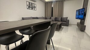 A modern living room with a dining area, a flat-screen TV mounted on the wall, and a ceiling light fixture. - JRE Cozy Home Near All Services 20 (Amman)