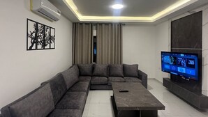 A modern living room with a sectional sofa, a coffee table, a flat-screen TV, and a wall-mounted air conditioner. - JRE Cozy Home Near All Services 20 (Amman)