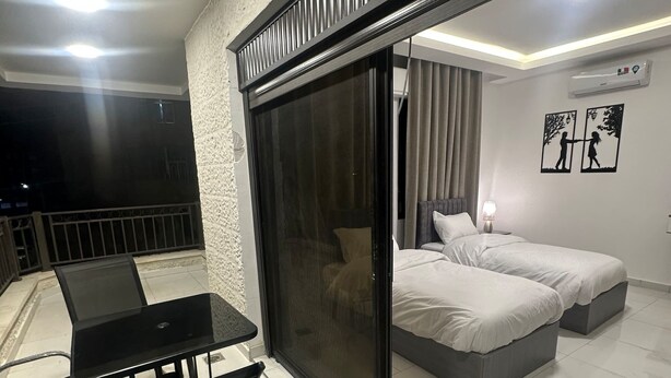 A modern room with a glass door leading to a balcony, a bed with two pillows, a small table with chairs, and a wall-mounted air conditioner. - JRE Cozy Home Near All Services 20 (Amman)