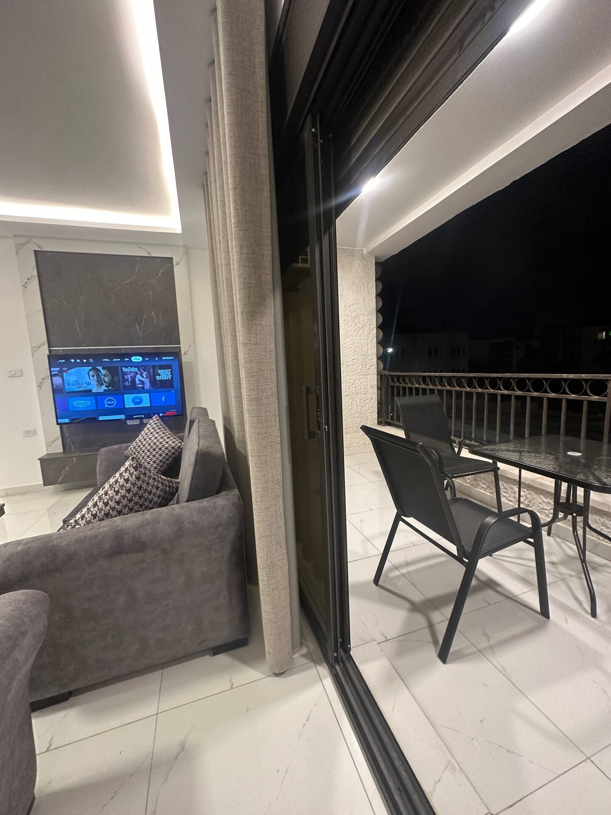 A modern living room with a sofa, a TV, and a glass door leading to an outdoor balcony with a table and chairs.
