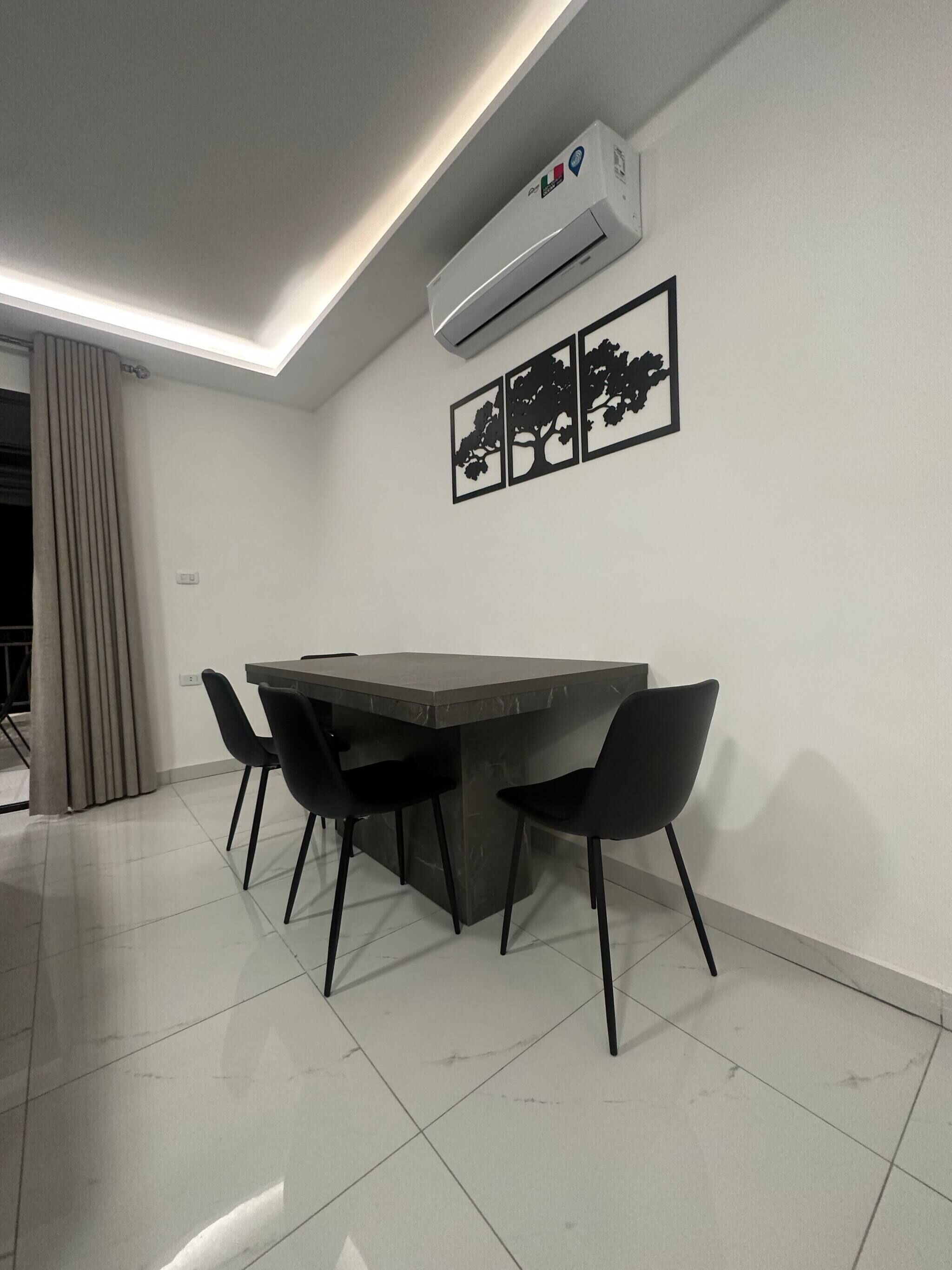 A modern dining area with a dark-toned table and four black chairs, an air conditioning unit, and three framed tree silhouettes on the wall.