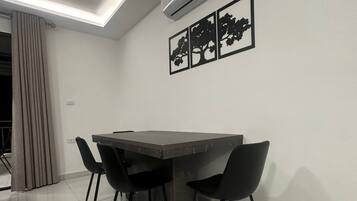 A modern dining area with a dark-toned table and four black chairs, an air conditioning unit, and three framed tree silhouettes on the wall.