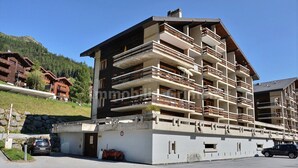 A multi-story hotel with balconies, a parking area, and a mountainous backdrop.