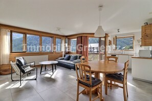 A spacious living area with a sofa, a dining table, and chairs. There is a kitchen area with cabinets and a microwave. Large windows provide a view of the outdoors. - Comfortable flat for 4 to 5 people, living room, kitchen area with dishwasher, 1 double bedroom, 1 3 (Grimentz)