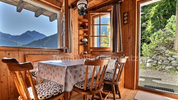 A wooden cabin with a round table and chairs, a window with a mountain view, and a shelf with kitchenware.