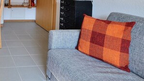 A modern kitchen with wooden chairs, a dining table, and a grey sofa with an orange pillow. - Comfortable flat for 2 to 4 people, living room with 1 double divan bed, kitchen area with dishwashe (Grimentz)