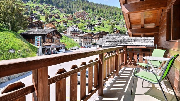 A balcony with wooden railings overlooking a village with traditional houses and a mountain backdrop.