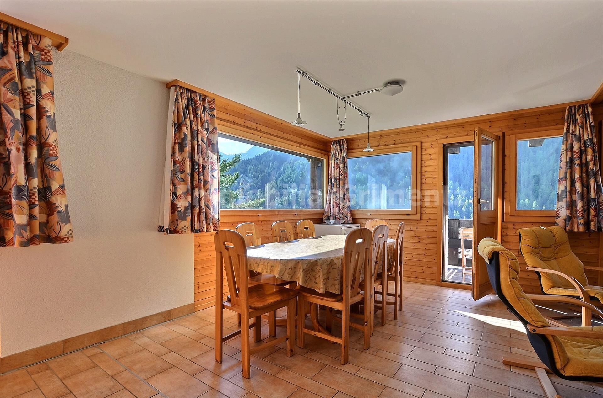 A wooden cabin with a dining table and chairs, large windows, and a ceiling light fixture.