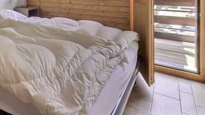 Een slaapkamer met een bed, een houten nachtkastje, een rode opbergla en een raam met jaloezieën.