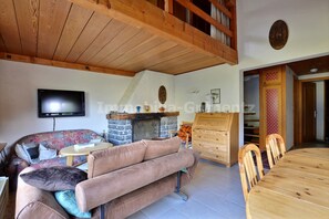 Uma sala de estar acolhedora com lareira de pedra, vigas de madeira no teto, uma televisão de tela plana, um sofá e uma mesa de jantar com cadeiras.