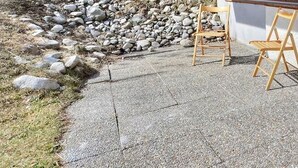 A stone-paved patio with two wooden chairs and a small table, a stone wall, and a bed inside a room. - Middle class flat for 5 people, living room with fireplace, kitchen area with dishwasher, 1 bedroom (Grimentz)