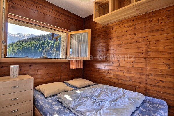 Une chambre rustique en bois, avec un lit, une commode et une fenêtre donnant sur une forêt et des montagnes.