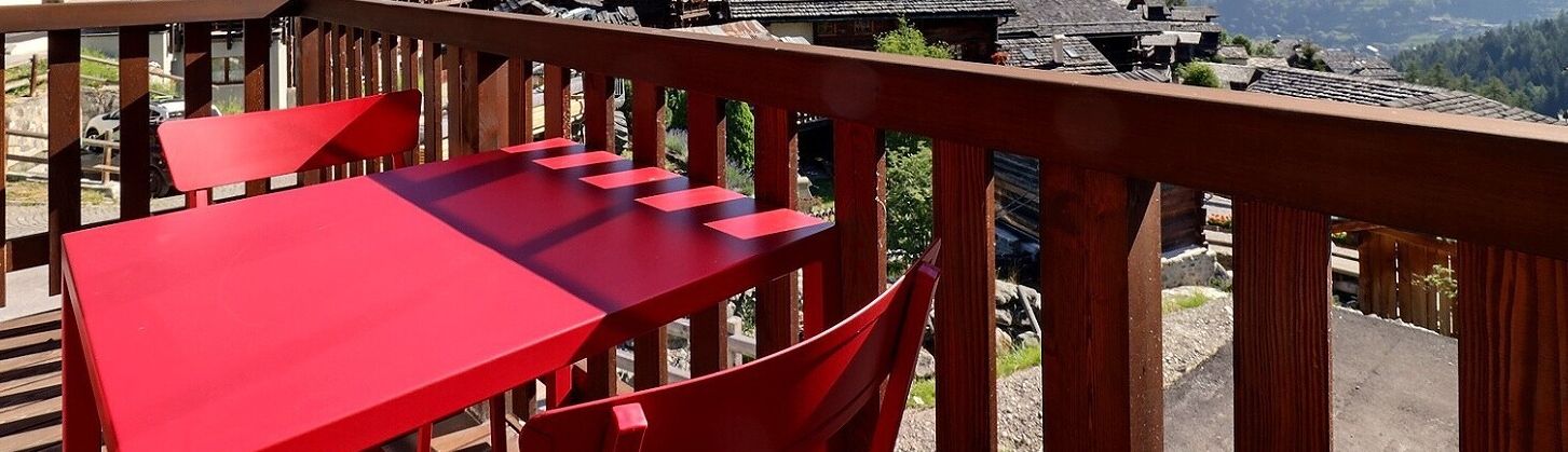Une table et une chaise rouges sur un balcon, avec vue sur des bâtiments traditionnels et une zone boisée.