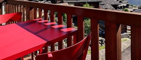 Une table et une chaise rouges sur un balcon, avec vue sur des bâtiments traditionnels et une zone boisée.