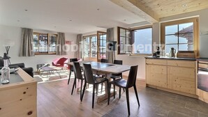 Un vaste espace de vie doté d’un plafond en bois, d’une table à manger avec des chaises, d’un coin cuisine avec des meubles en bois et de grandes fenêtres offrant une vue sur l’extérieur.