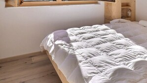 Un lit recouvert d’une couette blanche matelassée, dans une pièce au plancher en bois et avec une petite fenêtre au-dessus.