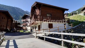 Un chalet en bois doté d’un balcon, un pont de pierre et une chaîne de montagnes en arrière-plan.