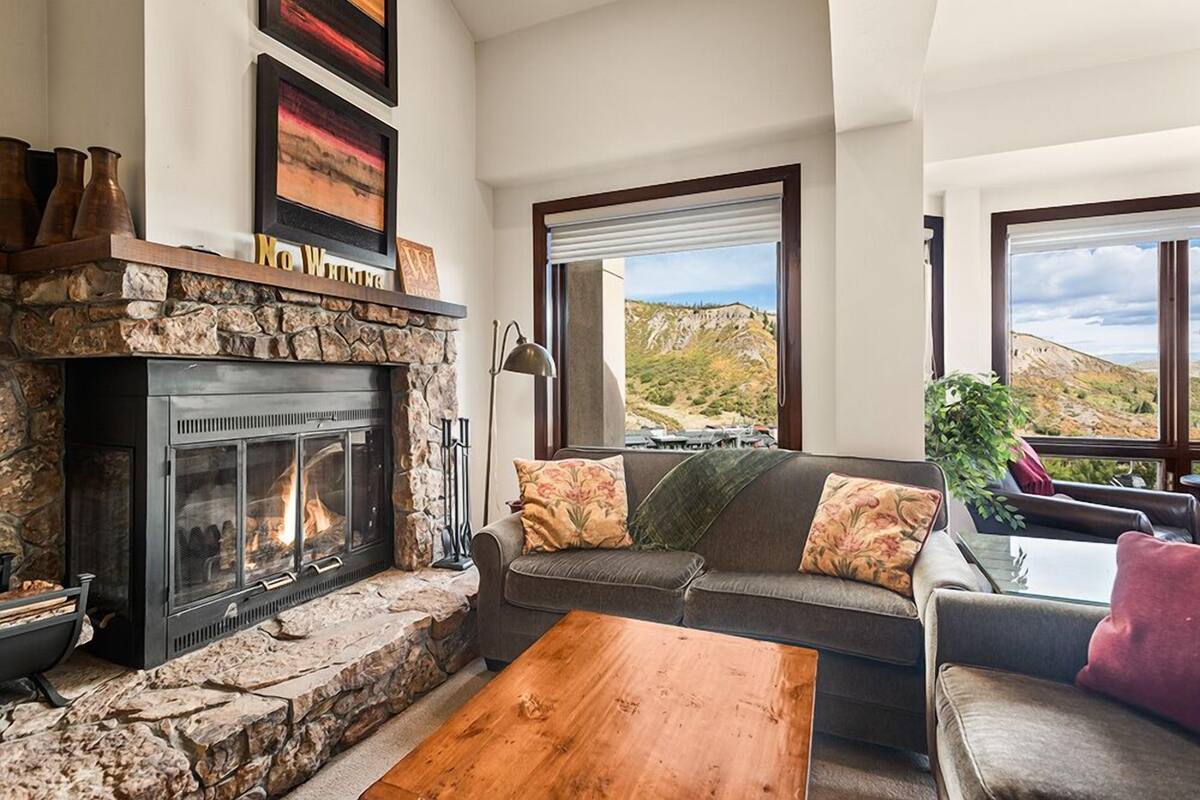 A cozy living room with a stone fireplace, a sofa with cushions, a wooden coffee table, and large windows offering a view of the outdoors.
