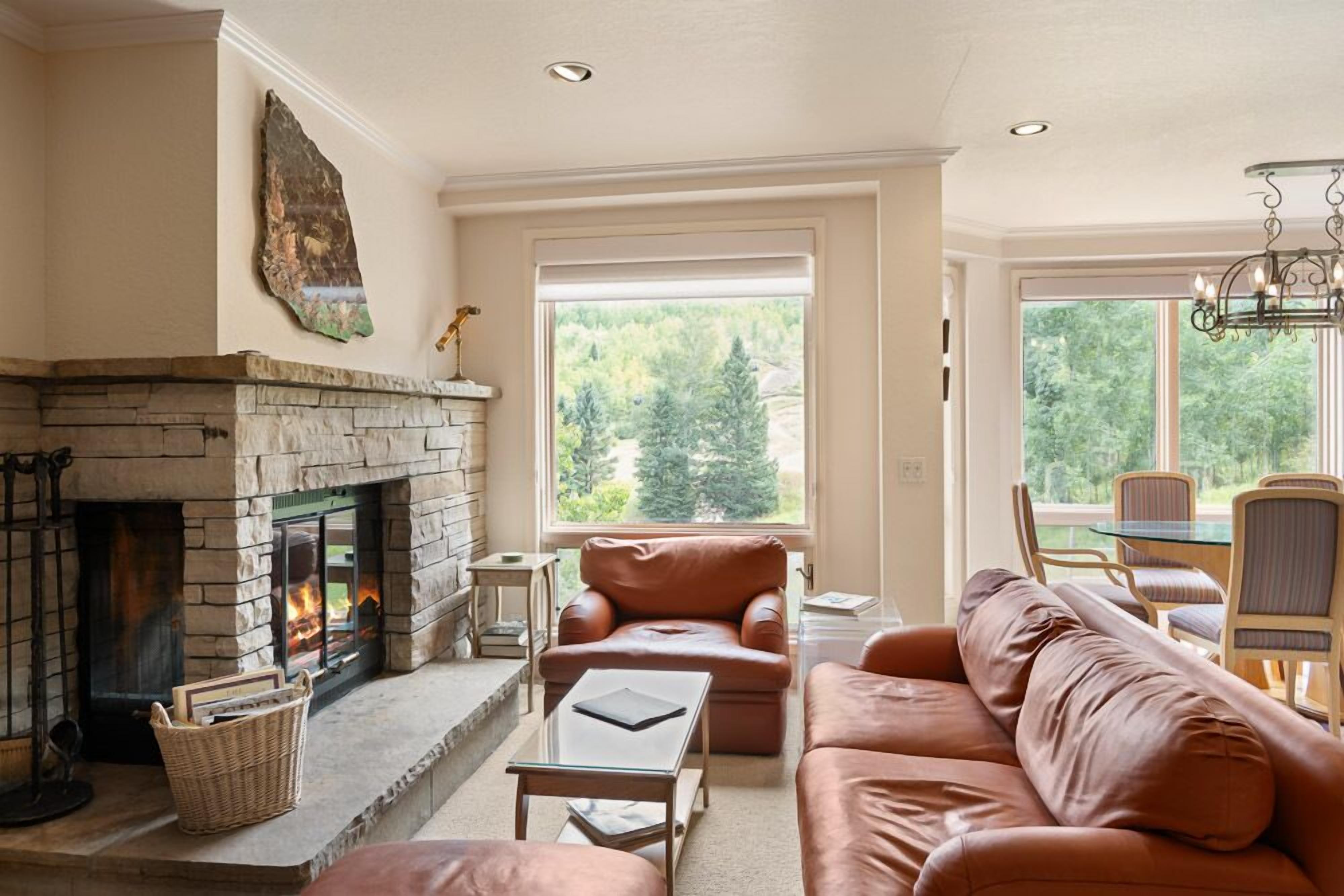 A living room with a stone fireplace, leather sofas, a glass coffee table, and large windows offering a view of trees.