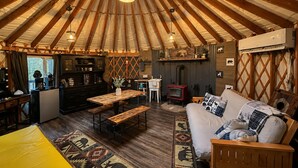 L’intérieur d’une yourte, avec une table en bois au centre, un canapé agrémenté de coussins à motifs, un petit coin cuisine et un luminaire suspendu.