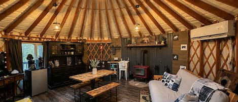 A yurt interior with a central wooden table, a sofa with patterned cushions, a small kitchen area, and a hanging light fixture.