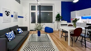 Ein modernes Wohnzimmer mit einer Couch, einem Couchtisch und einem Essbereich mit Stühlen und einem Tisch. Ein Fenster mit Jalousien, eine Pflanze und Kunstwerke schmücken die Wände.