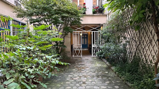 Ein schmaler Pflasterweg führt zu einem verschlossenen Eingang, umgeben von üppigem Grün und einem Gebäude mit Balkon.