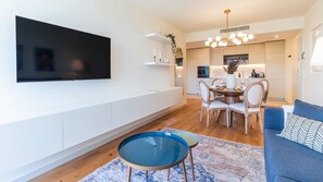 A modern living room with a flat-screen TV, a dining area with a round table and chairs, and a kitchen in the background.