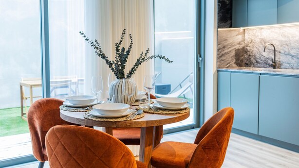 A dining table set with plates, glasses, and a vase with greenery, in a room with large windows, a kitchen area, and orange chairs. - RUAH C - Cozy Studio by LovelyStay (Vila Nova de Gaia)