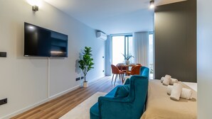 A modern hotel room with a flat-screen TV mounted on the wall, a teal sofa, a dining area with a table and chairs, and a large window with curtains. - RUAH C - Cozy Studio by LovelyStay (Vila Nova de Gaia)