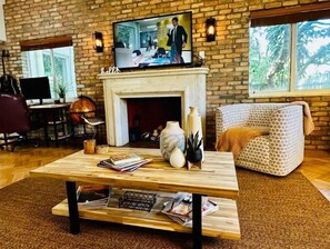 Una sala de estar acogedora con chimenea, un televisor montado encima, un sofá estampado y una mesa de centro de madera con libros y objetos decorativos.
