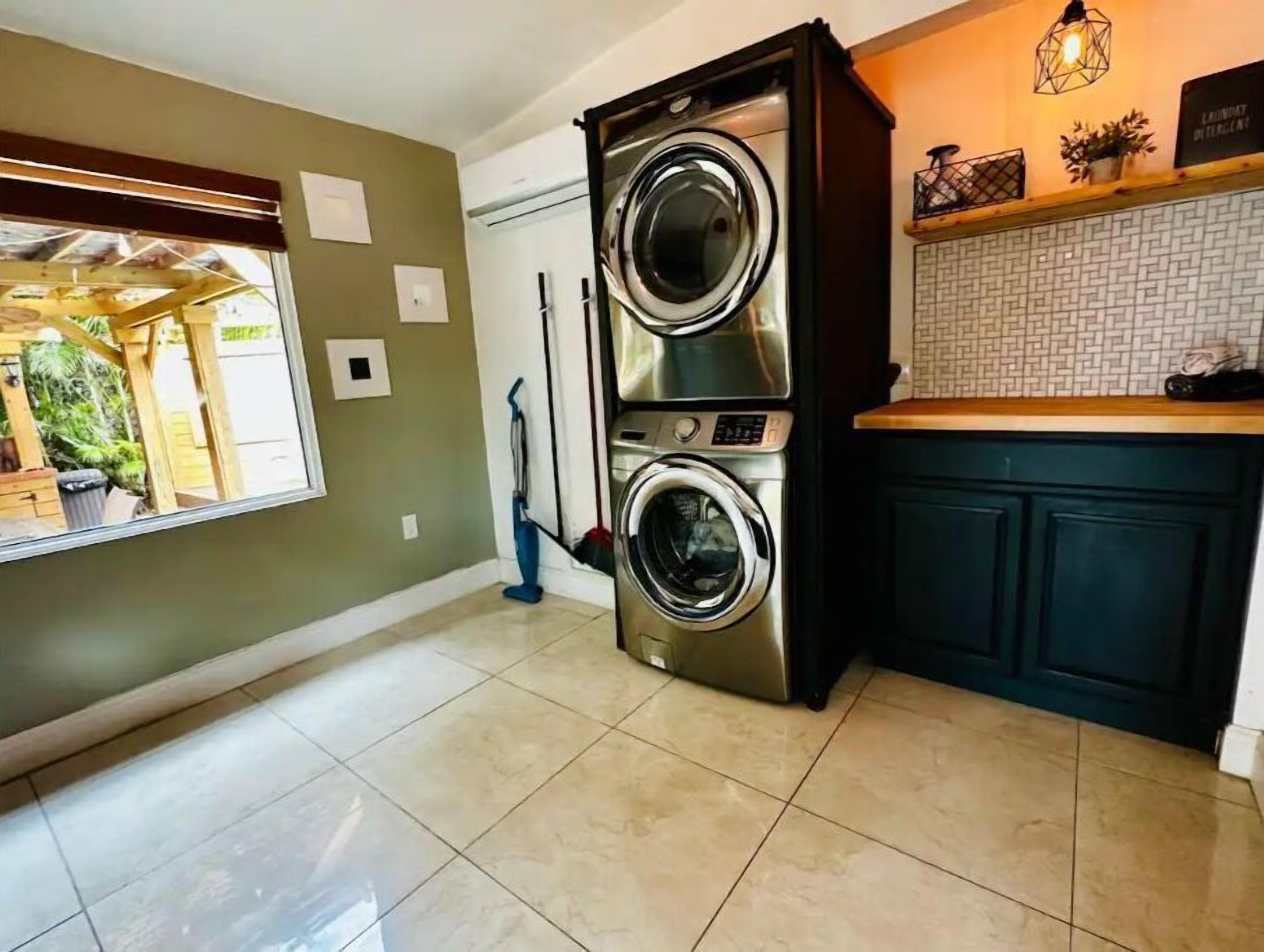 A laundry room with a stacked washer and dryer, a mop, and a window with a view of a garden.
