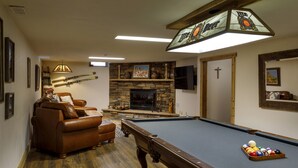 A pool table with a billiard ball rack, a leather sofa, a stone fireplace, and a cross on the wall.