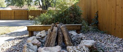 Bocche di fuoco circondata da rocce e tronchi, con una panca di legno e un recinto in legno sullo sfondo.