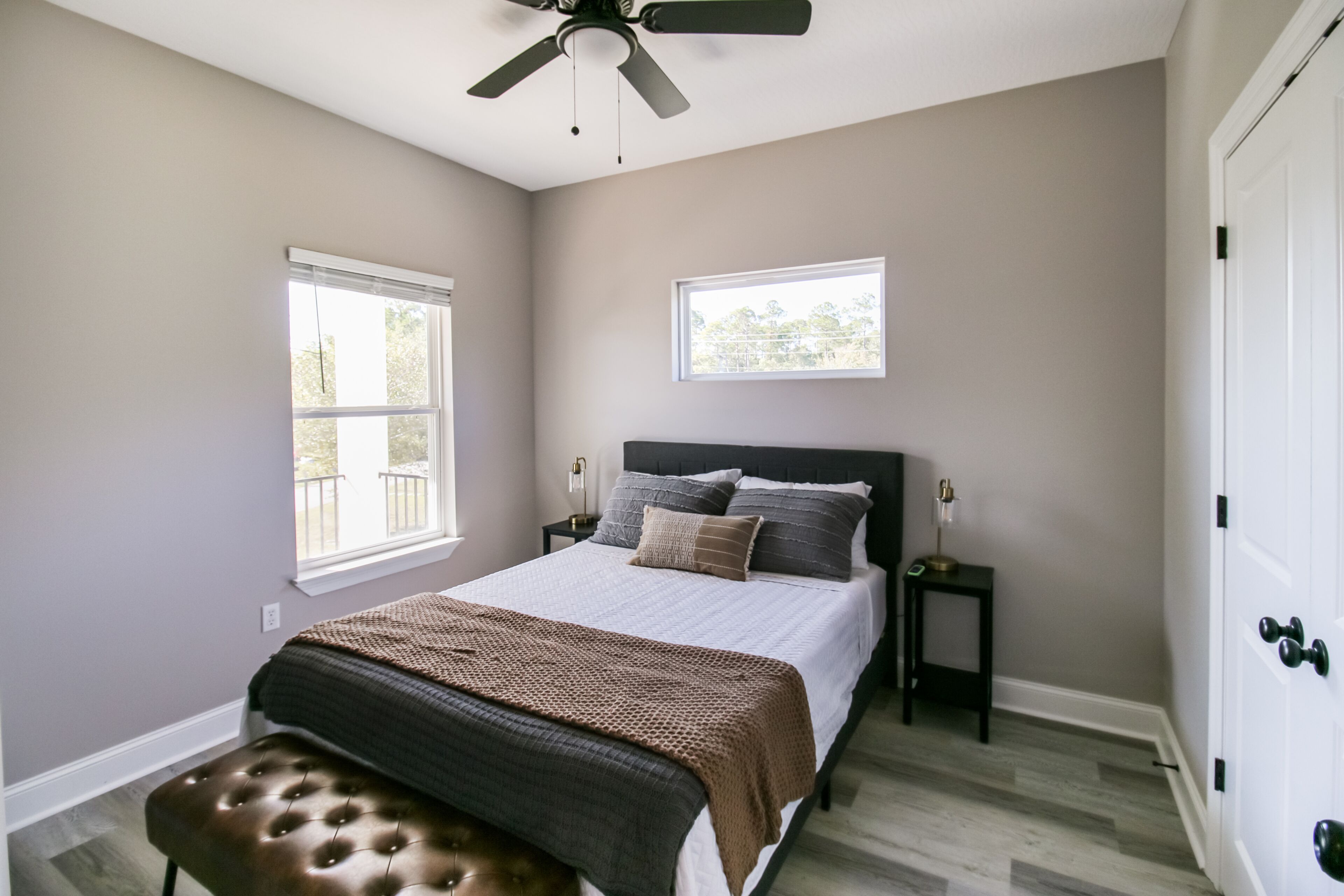 A bedroom with a bed, two bedside tables, a bench, a ceiling fan, and two windows.