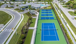 An aerial view of a tennis court complex with multiple courts, a parking area, and surrounding greenery.