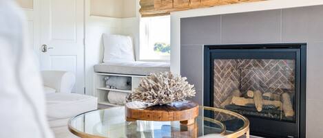 A living room with a fireplace, a white sofa, a glass coffee table, and a wooden shelf above the fireplace.