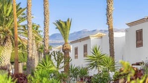 Une rangée de bâtiments blancs aux toits de tuiles rouges, entourés de palmiers et de cactus, avec une chaîne de montagnes en arrière-plan.