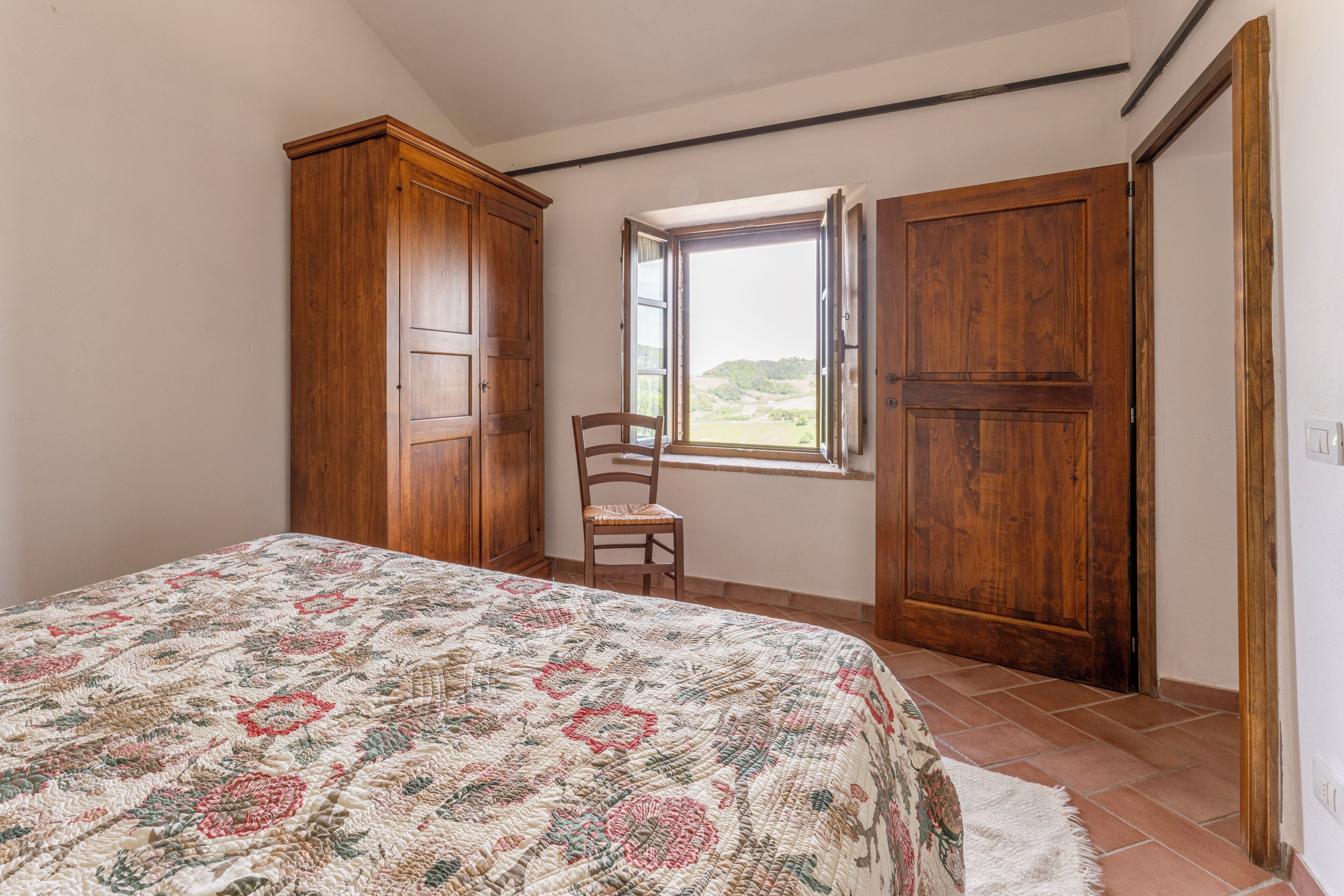 Ein Schlafzimmer mit einem Bett, einem Holzschrank, einem Stuhl und einem Fenster mit Blick ins Freie.